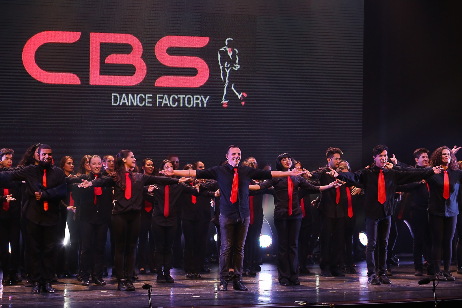 Bailarinos da CBS no palco, com figurinos pretos e gravatas vermelhas